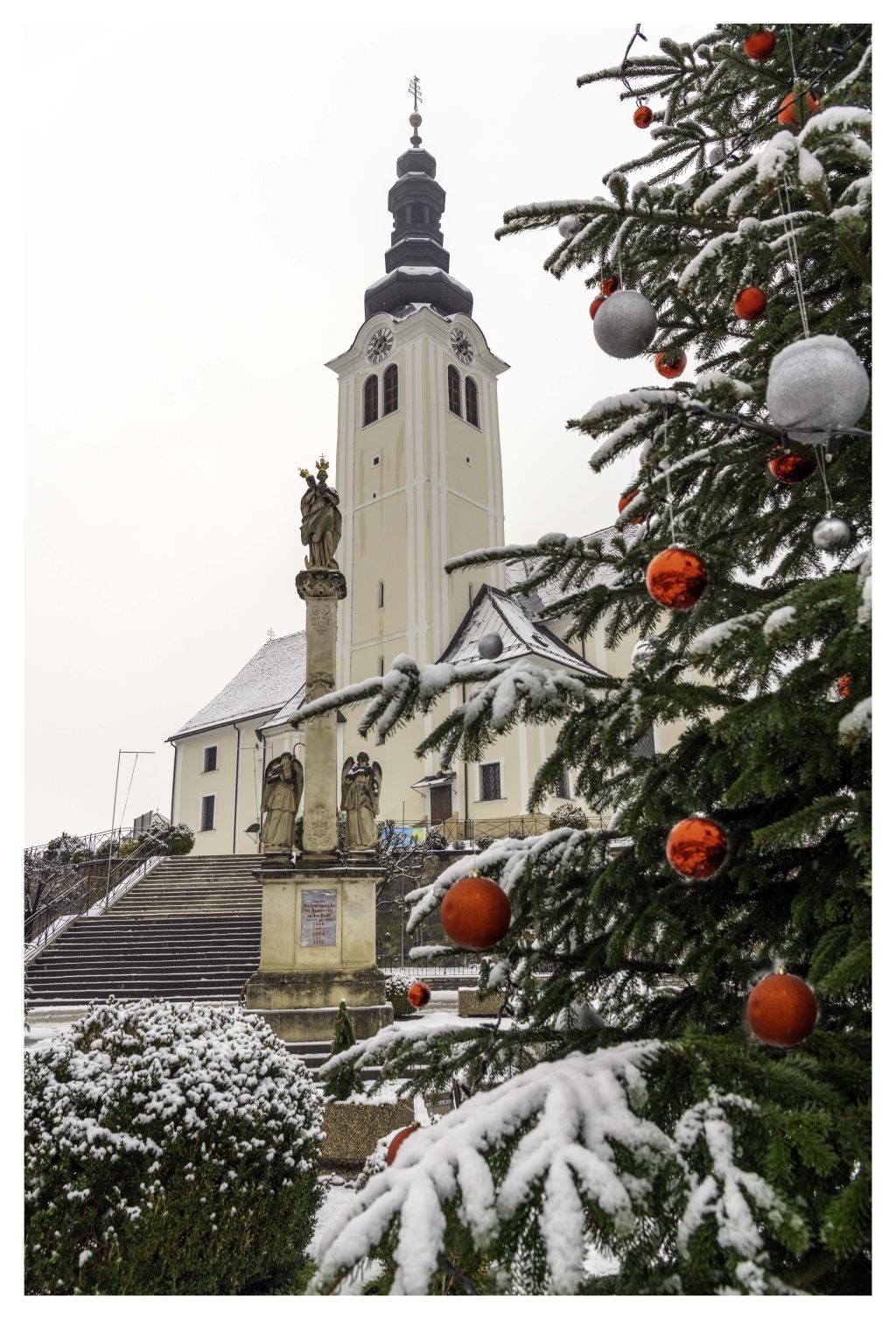 Eine Kirche mit einem hohen Turm und einer Statue eines Engels davor, umgeben von Schnee und einem Weihnachtsbaum mit roten und silbernen Ornamenten.