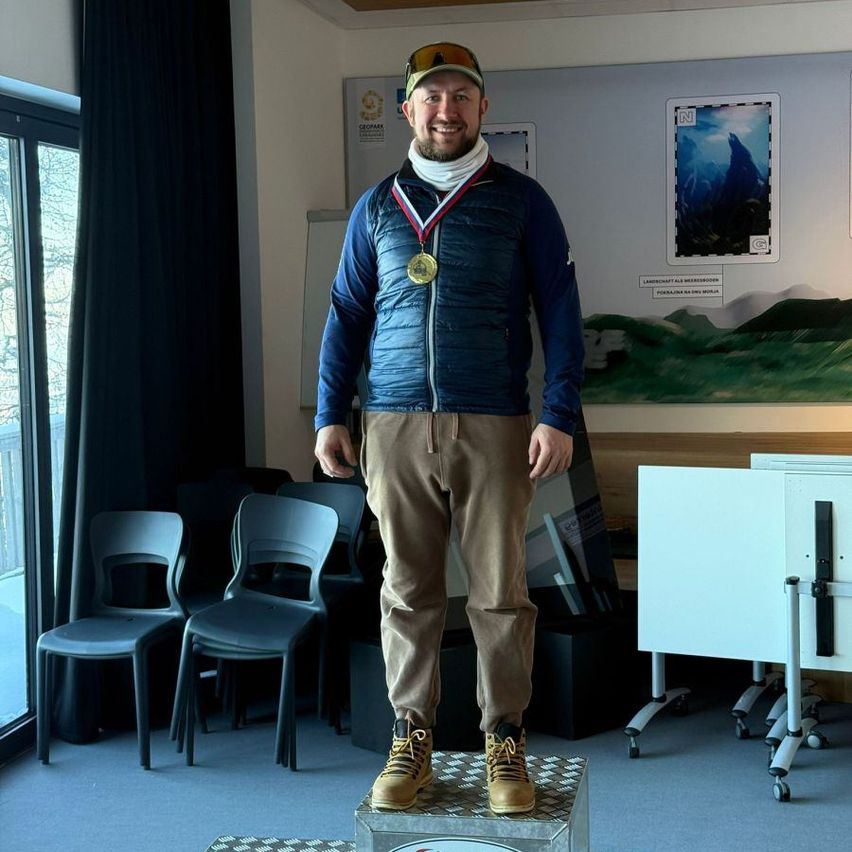 A man stands on a podium wearing a medal around his neck. He is wearing a blue jacket, brown pants, and sneakers. Behind him, there are chairs, a whiteboard, and a large wall with pictures.