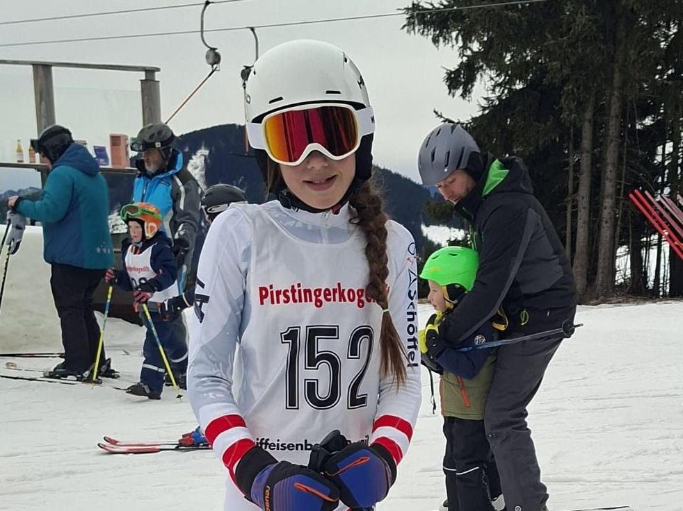 A young girl with braided hair and goggles stands on a snowy slope. She wears a white jersey with the number 152. Behind her, a man holds a child in a green helmet. Several other children and adults are in the background.