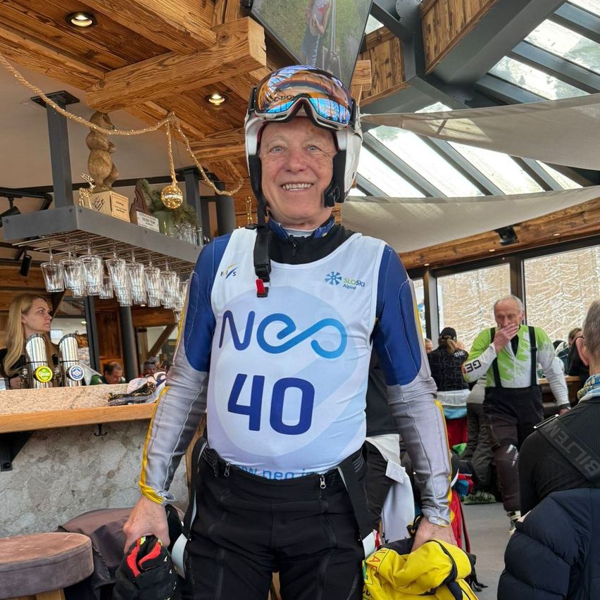 A man wearing a helmet and ski gear stands in a bar, smiling and holding ski equipment. Behind him, a woman tends to a bar, with shelves holding glasses and trophies. Other patrons are present, and a mountain view is visible through the glass walls.