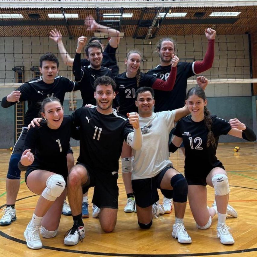 A group of volleyball players poses for a photo on the court, with numbers on their jerseys. The team is wearing black uniforms.