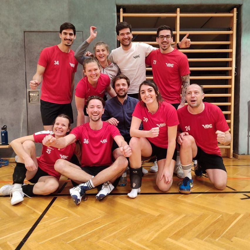 A group of people wearing red jerseys with numbers pose for a photo on a wooden floor. They are smiling and posing for the camera. Behind them is a wooden structure.