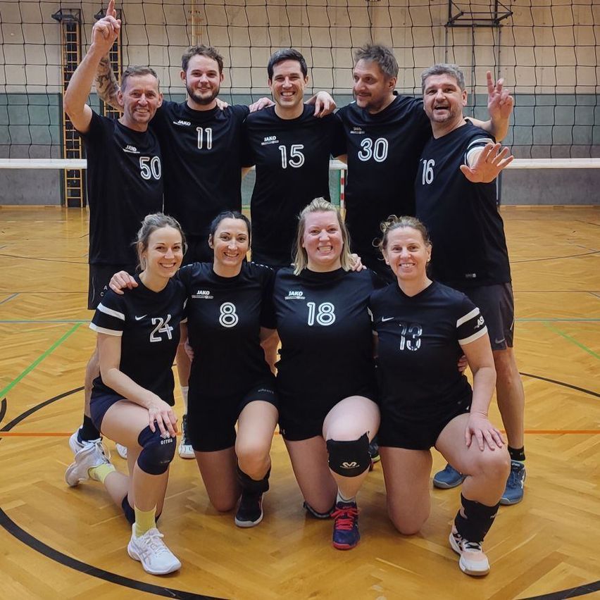 A group of volleyball players in black jerseys, with numbers ranging from 8 to 50, are posing for a photo on an indoor court.