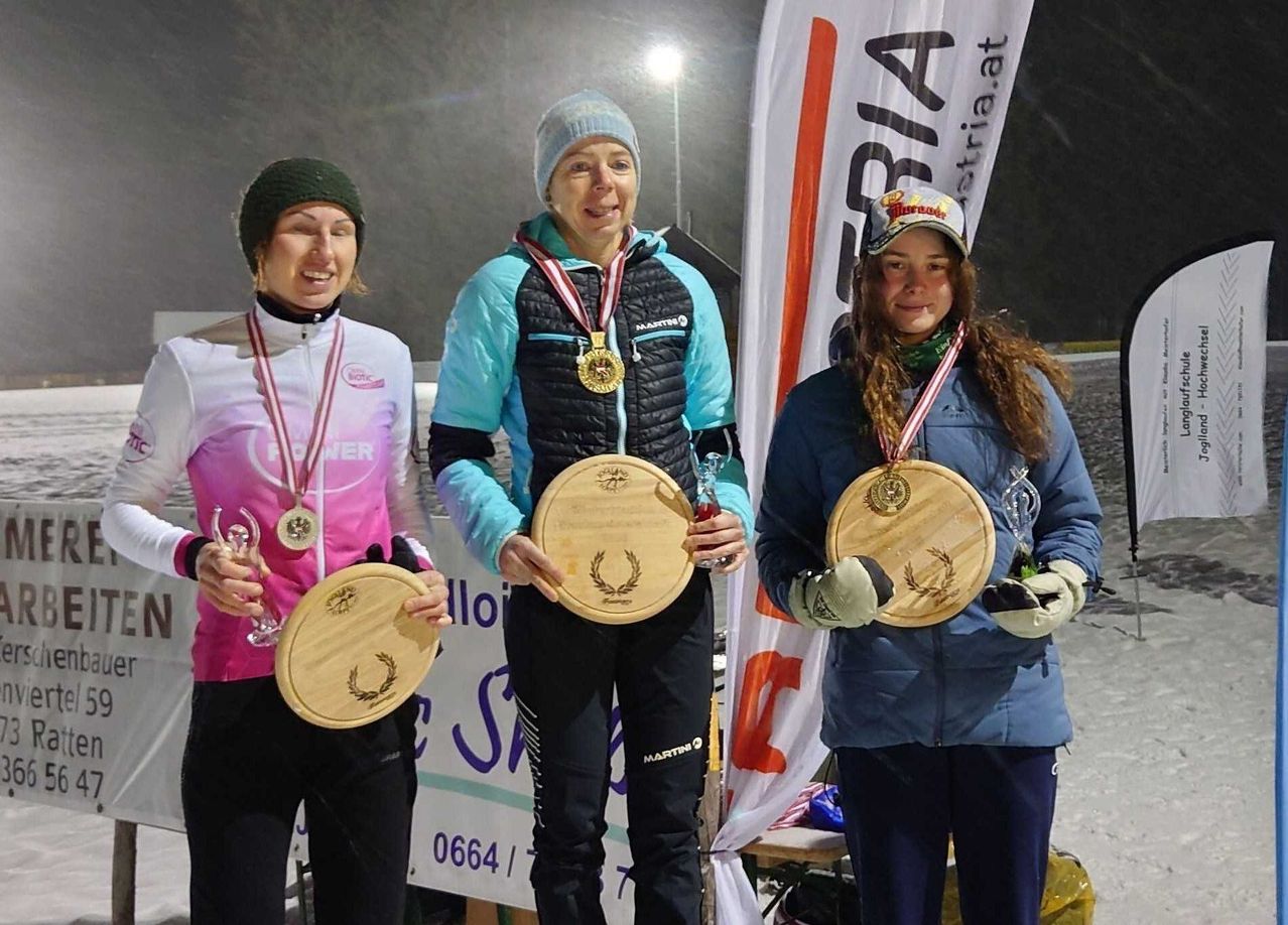 Drei Frauen posieren bei Nacht für ein Foto, tragen Medaillen und halten Holzplaketten. Sie lächeln, eine hält ein Glas. Ein Banner mit dem Wort 'AUSTRIA' steht hinter ihnen.