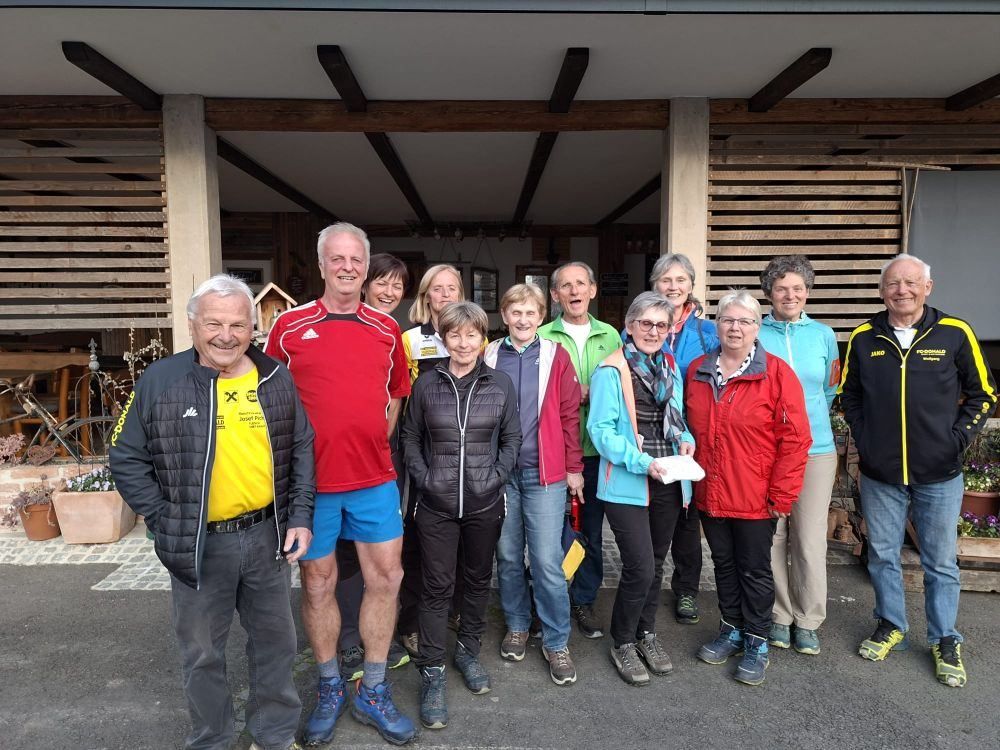 A group of smiling people, dressed in various colored jackets, stands together in front of a wooden building.