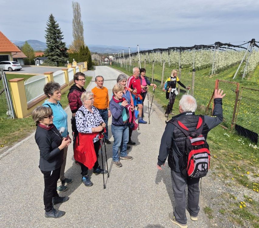 A group of people, some with hiking gear, stands on a gravel path with a fence, orchard, and houses in the background.