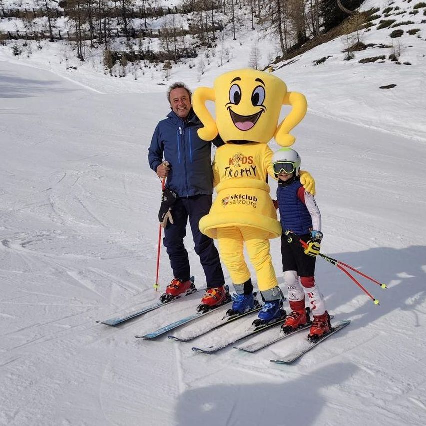Ein Mann und ein Kind fahren auf einer verschneiten Piste Ski, begleitet von einem Maskottchen in einem gelben Pokalkostüm.