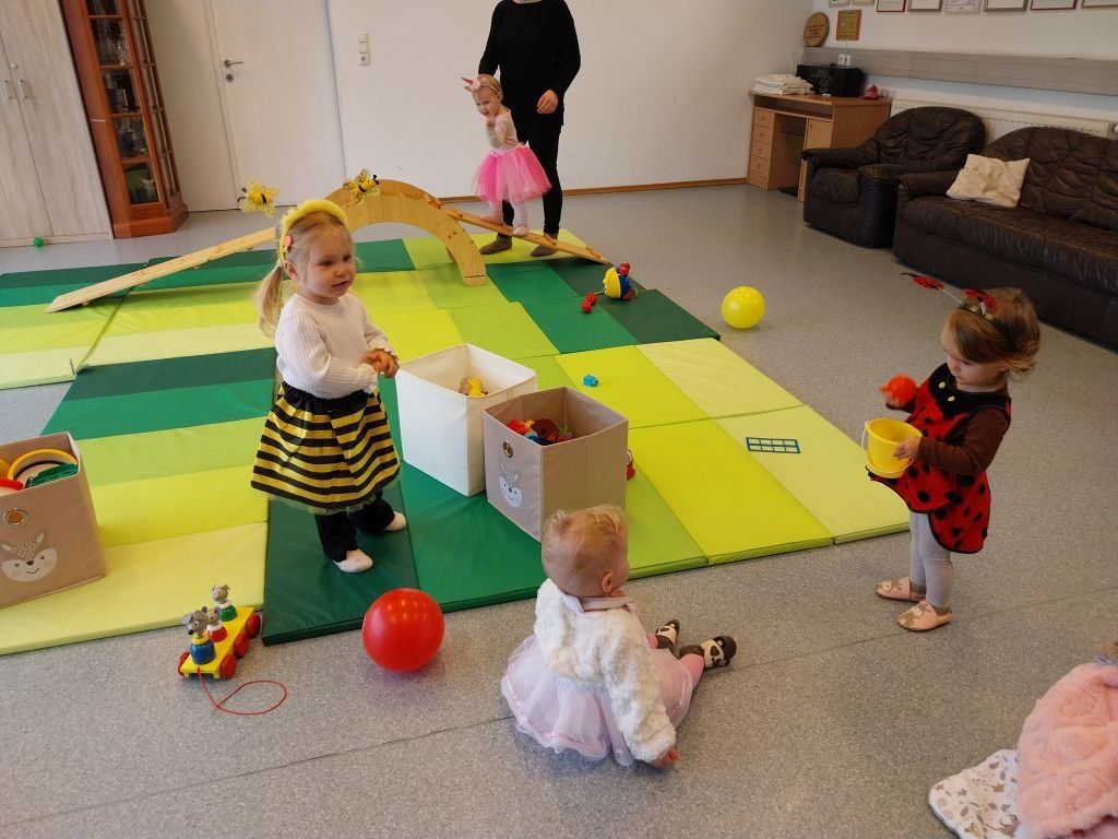 Kinder spielen auf einem bunten Teppich mit Spielzeug. Ein Kleinkind sitzt auf dem Boden und ein Mädchen in Bienenkostüm steht in der Nähe. Ein Erwachsener steht hinter ihnen.
