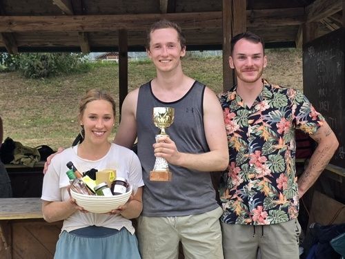 Three people stand together outside, one holding a trophy and a bowl of items, smiling for a photo. Behind them is a wooden structure with a blackboard.