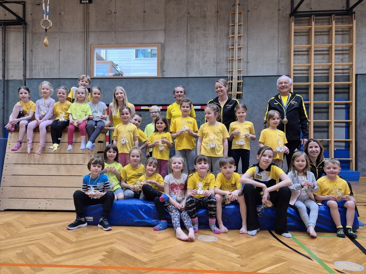 Eine Gruppe von Kindern in gelben Shirts posiert mit ihren Trainern in einer Turnhalle. Sie halten Medaillen und einige sitzen auf einer blauen Matte.