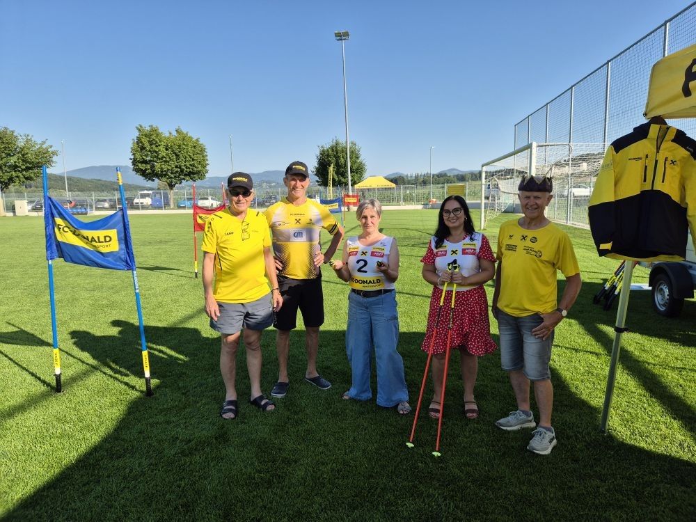 Fünf Personen stehen auf einem Rasenfeld, wahrscheinlich bei einer Veranstaltung, und tragen gelbe T-Shirts. Eine Frau hält ein Schild mit der Nummer 2, und eine andere hält Skistöcke. Hinter ihnen sind ein Zaun, Bäume und Berge zu sehen.