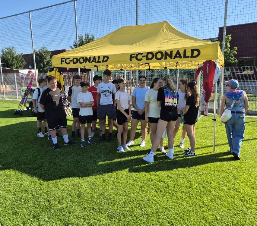 Eine Gruppe von Schülern steht unter einem gelben Zelt namens FC-DONALD, einige mit Rucksäcken und Sportausrüstung. Sie sind von einem Zaun und Bäumen umgeben.