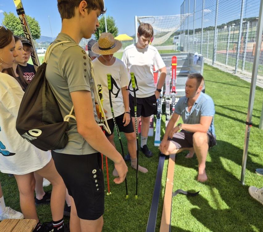 Eine Gruppe von Menschen versammelt sich im Freien. Ein Mann kniet und untersucht Skiausrüstung. Ein junger Mann sieht zu, während andere in der Nähe zusehen.