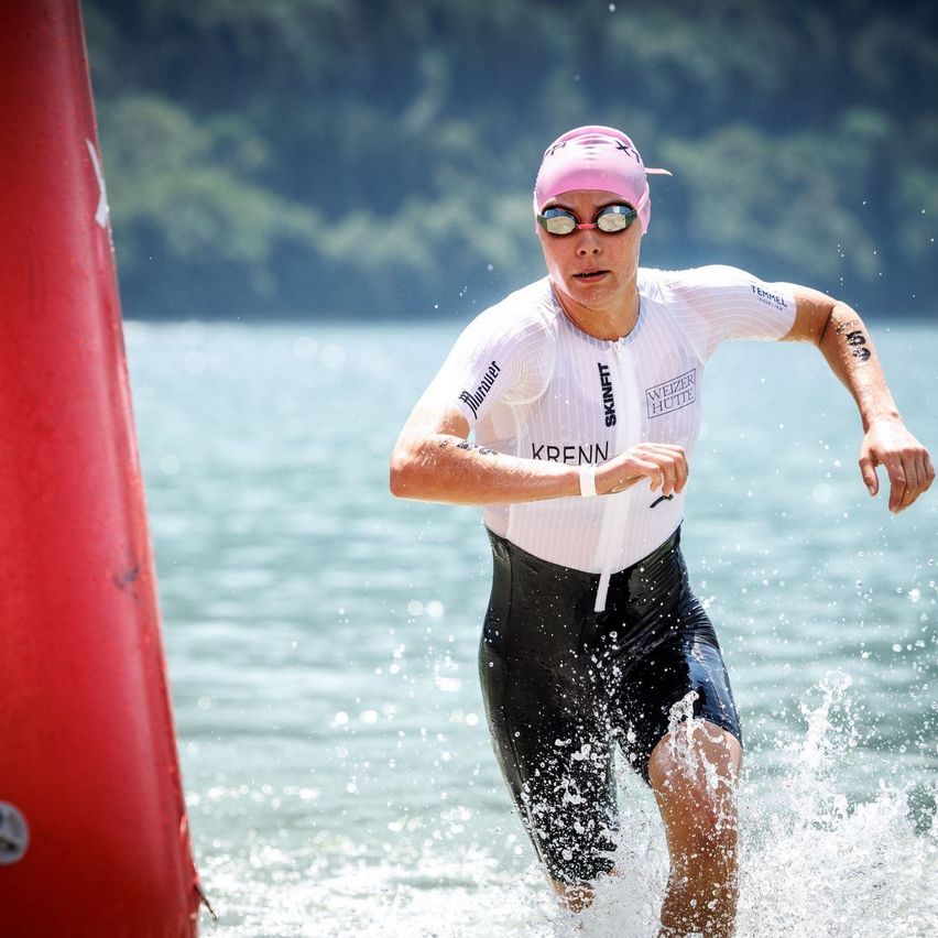 Eine Triathletin verlässt das Wasser und rennt auf einen roten Marker zu. Sie trägt einen weißen und schwarzen Rennanzug, eine pinke Kappe und eine Schwimmbrille.