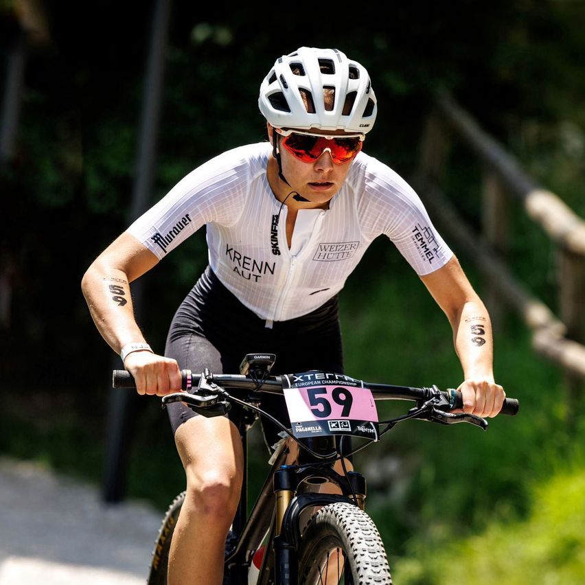 Eine Radfahrerin fährt bergab, trägt einen weißen Helm, eine Sonnenbrille und ein weißes Trikot mit 'Krenn Aut' und 'Weizer Hutte' Aufdruck. Die Nummer '59' ist am Lenker.