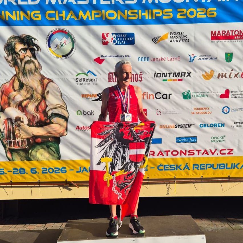 Ein Mann steht auf einem Podest mit Medaillen und hält eine österreichische Flagge in der Hand. Er befindet sich bei den World Masters Athletics Championships 2026. Hinter ihm ist ein großes Banner.