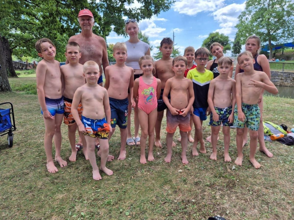 A group of children and adults stand together on a grassy field, smiling for a photo. The children wear swimsuits, and the adults include a man with a red cap and a woman with sunglasses on her head. Trees and a cloudy sky are in the background.