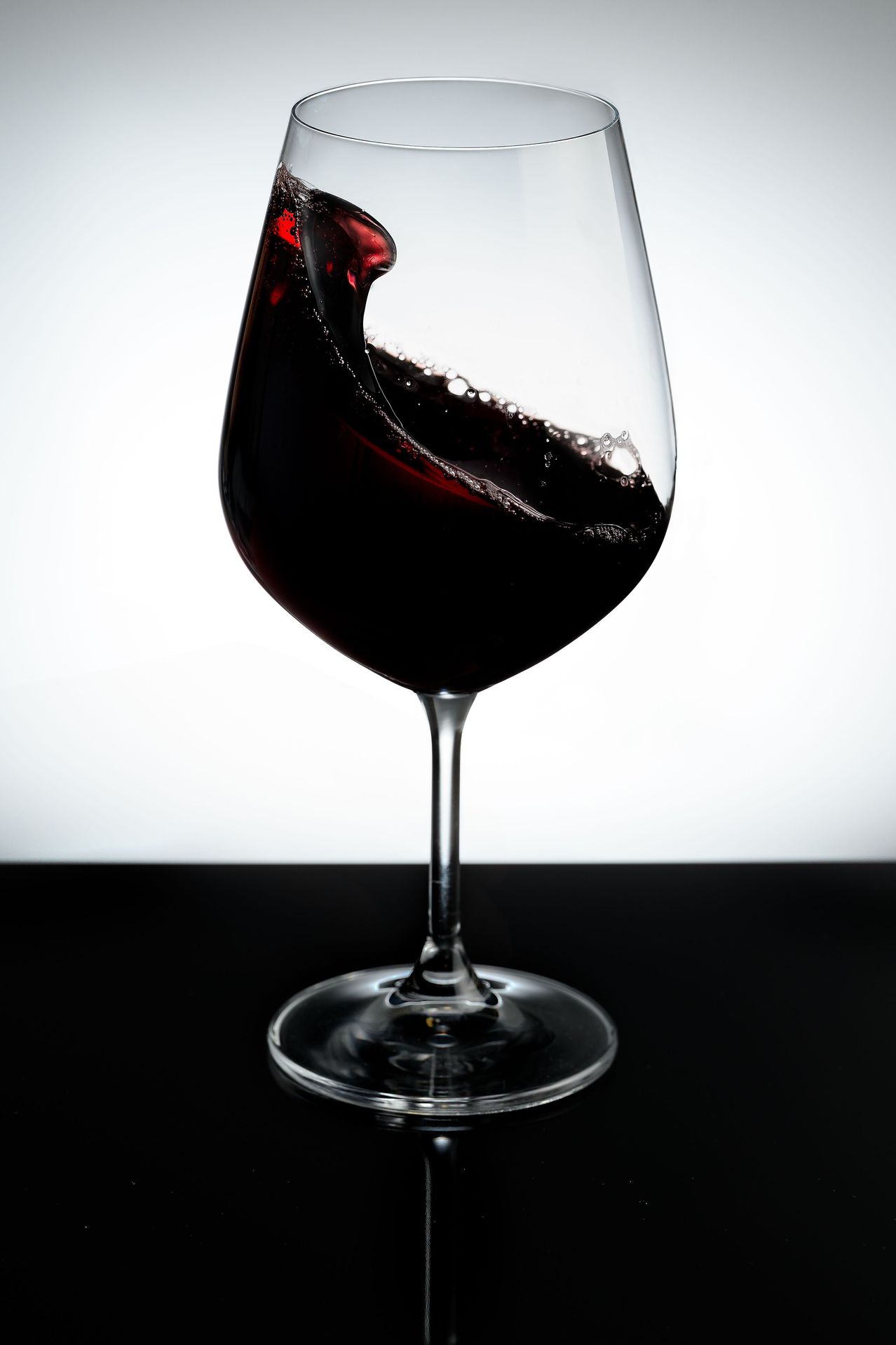 Ein Glas Rotwein mit einer tiefen, reichen Farbe, auf einem schwarzen Tisch vor weißem Hintergrund platziert.