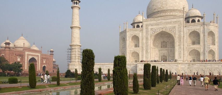 Der Taj Mahal ist ein großes weißes Marmormausoleum mit einer Kuppel, vier Minaretten und kunstvollen Schnitzereien. Besucher gehen auf einem gepflasterten Weg vor dem Gebäude entlang.