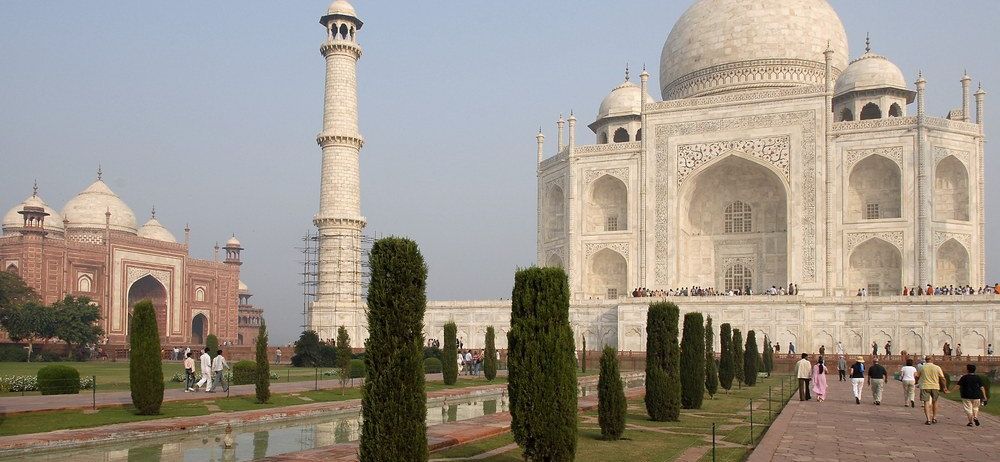 Der Taj Mahal ist ein großes weißes Marmormausoleum mit einer Kuppel, vier Minaretten und kunstvollen Schnitzereien. Besucher gehen auf einem gepflasterten Weg vor dem Gebäude entlang.