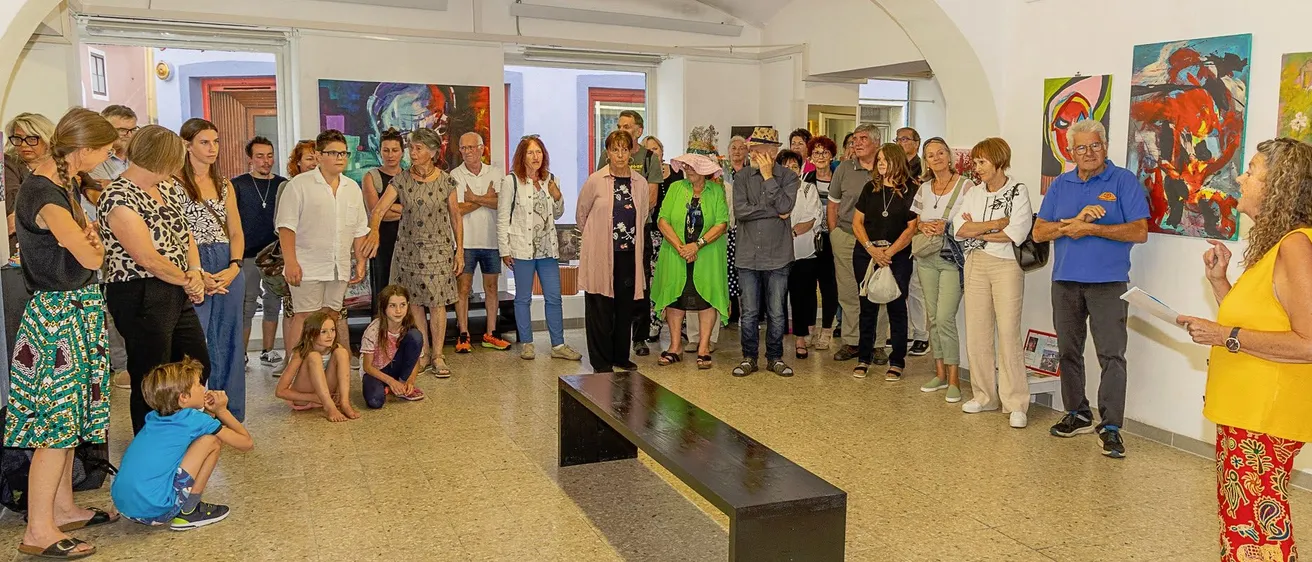 Eine Gruppe von Menschen ist drinnen versammelt, wahrscheinlich in einer Kunstgalerie. Sie betrachten Gemälde an der Wand. Einige von ihnen stehen, während andere auf dem Boden sitzen. Eine lange Bank steht in der Mitte des Raumes.