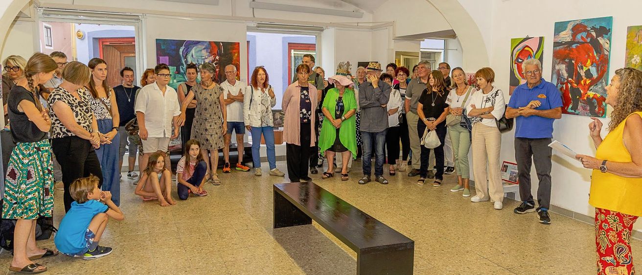 Eine Gruppe von Menschen ist drinnen versammelt, wahrscheinlich in einer Kunstgalerie. Sie betrachten Gemälde an der Wand. Einige von ihnen stehen, während andere auf dem Boden sitzen. Eine lange Bank steht in der Mitte des Raumes.