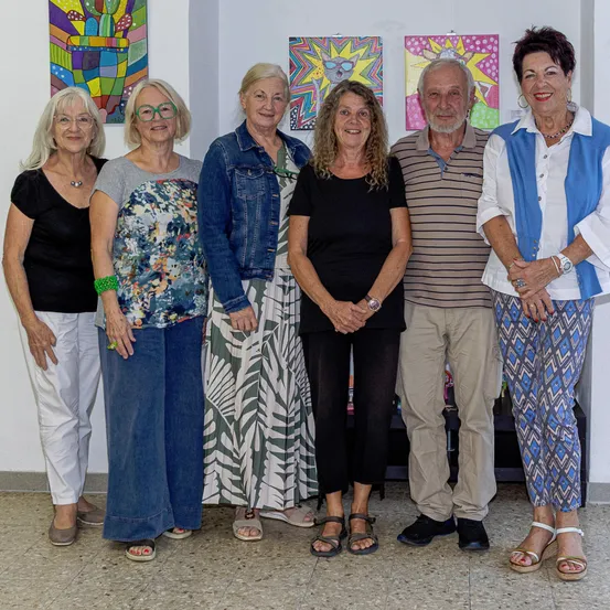 Eine Gruppe von sechs Erwachsenen, darunter drei Frauen und drei Männer, steht gemeinsam vor einer Wand, die mit farbenfrohen Gemälden geschmückt ist. Sie alle lächeln und scheinen für ein Foto zu posieren.