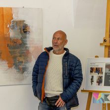 Ein Mann steht vor einem modernen abstrakten Gemälde, trägt eine blaue Jacke und ein weißes Hemd und schaut zur Kamera.