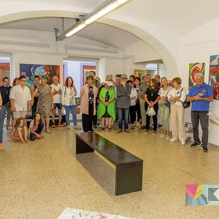 Eine Gruppe von Menschen versammelt sich in einer Kunstgalerie, betrachtet Gemälde an den Wänden und führt Gespräche. Eine schwarze Bank steht im Mittelpunkt, und einige Personen sitzen oder knien auf dem Boden.