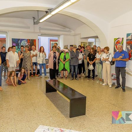 Eine Gruppe von Menschen versammelt sich in einer Kunstgalerie, betrachtet Gemälde an den Wänden und führt Gespräche. Eine schwarze Bank steht im Mittelpunkt, und einige Personen sitzen oder knien auf dem Boden.