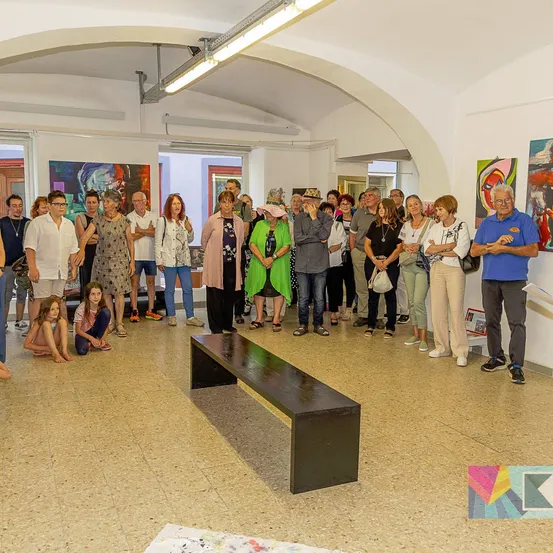 Eine Gruppe von Menschen versammelt sich in einer Kunstgalerie, betrachtet Gemälde an den Wänden und führt Gespräche. Eine schwarze Bank steht im Mittelpunkt, und einige Personen sitzen oder knien auf dem Boden.