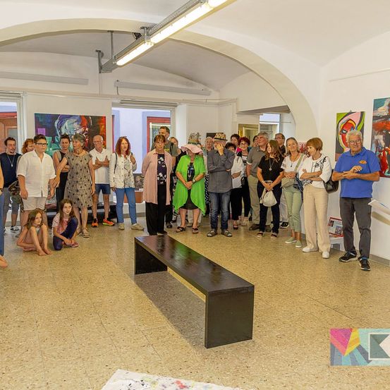 Eine Gruppe von Menschen versammelt sich in einer Kunstgalerie, betrachtet Gemälde an den Wänden und führt Gespräche. Eine schwarze Bank steht im Mittelpunkt, und einige Personen sitzen oder knien auf dem Boden.