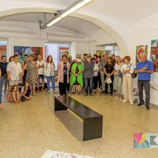 Eine Gruppe von Menschen versammelt sich in einer Kunstgalerie, betrachtet Gemälde an den Wänden und führt Gespräche. Eine schwarze Bank steht im Mittelpunkt, und einige Personen sitzen oder knien auf dem Boden.