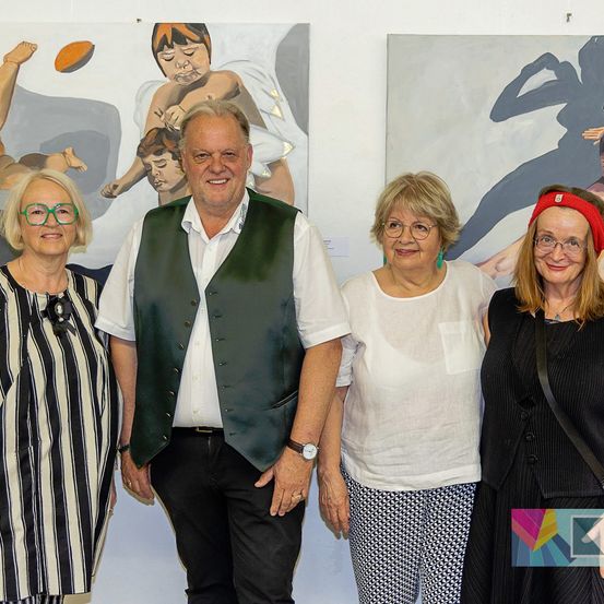 Fünf Erwachsene stehen vor zwei großen Gemälden an einer Wand und lächeln für ein Foto. Der Mann in der Mitte trägt eine grüne Weste.