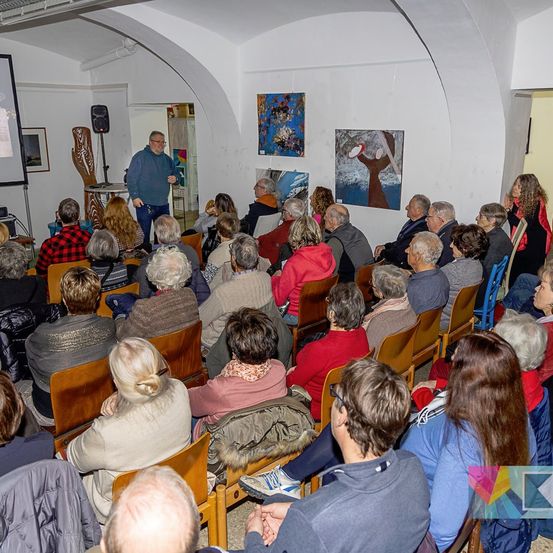 Ein Mann steht vor einer Projektionsleinwand und spricht eine sitzende Zuhörerschaft an. Der Raum hat gewölbte Decken und Gemälde an den Wänden. Die Zuhörerschaft ist vielfältig in Alter und Kleidung.