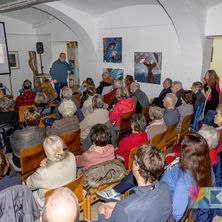 Ein Mann steht vor einer Projektionsleinwand und spricht eine sitzende Zuhörerschaft an. Der Raum hat gewölbte Decken und Gemälde an den Wänden. Die Zuhörerschaft ist vielfältig in Alter und Kleidung.