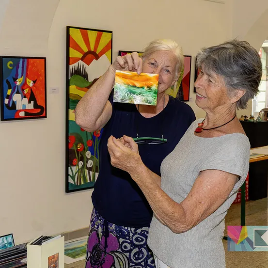 Zwei ältere Frauen stehen in einer Kunstgalerie, eine hält ein Gemälde hoch und die andere schaut es sich an. Die Galerie hat Gemälde an der Wand.