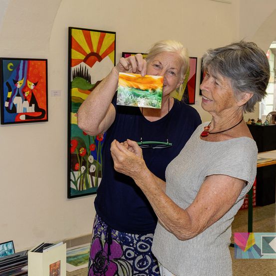 Zwei ältere Frauen stehen in einer Kunstgalerie, eine hält ein Gemälde hoch und die andere schaut es sich an. Die Galerie hat Gemälde an der Wand.