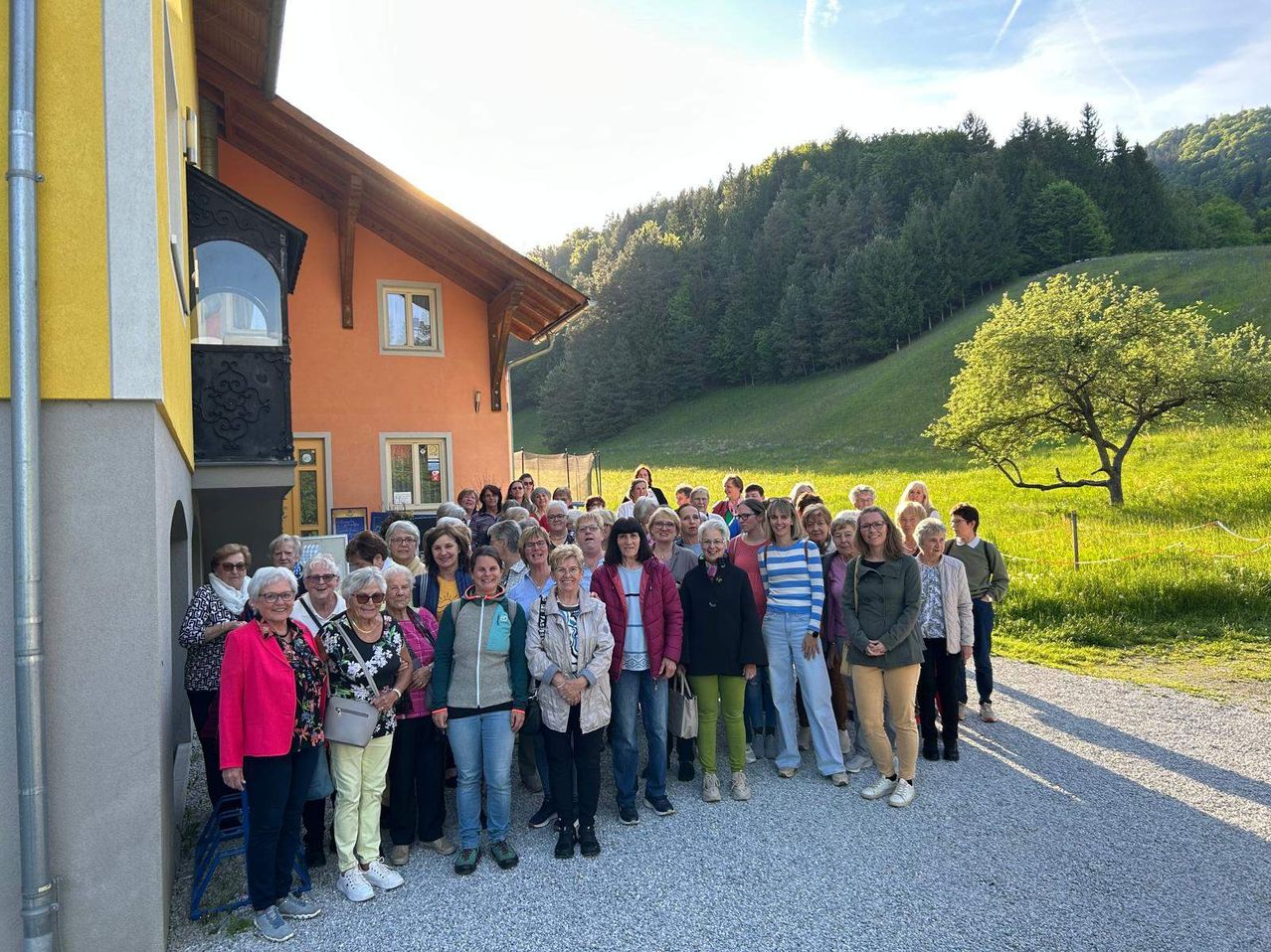 Eine Gruppe von Menschen in lässiger Kleidung versammelt sich vor einem Gebäude, wahrscheinlich einem Haus, an einem sonnigen Tag. Im Hintergrund befindet sich ein grasbewachsener Hügel mit Bäumen.