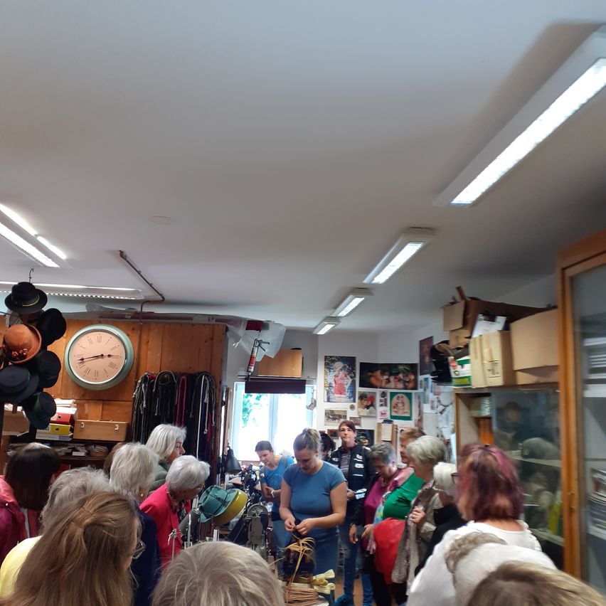 Eine Menschenmenge versammelt sich in einem Raum mit Holzwänden und an der Decke hängenden Hüten. Eine Frau in einem blauen Shirt steht in der Mitte und hält einen Hut. Verschiedene Gegenstände sind auf Regalen ausgestellt, und eine Uhr hängt an der Wand.