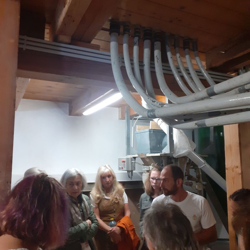 Eine Gruppe von Menschen steht in einem Raum mit weißen Rohren an der Decke. Ein Mann mit Bart spricht. Die anderen hören zu. Eine Maschine ist im Hintergrund.