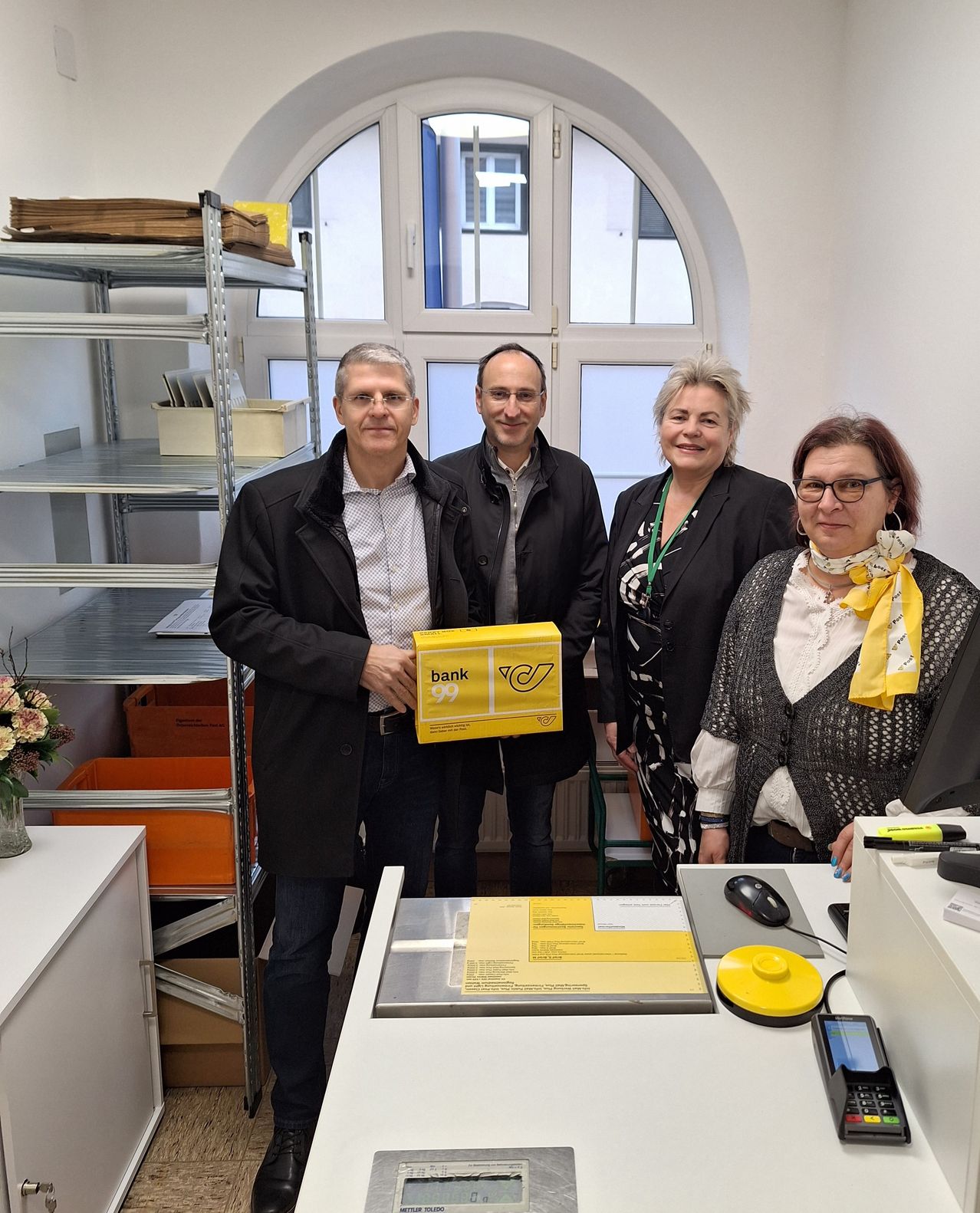 Vier Personen stehen lächelnd in einem Büro, einer von ihnen hält eine gelbe Box. Hinter ihnen ist ein Regal mit Gegenständen und ein Fenster zu sehen. Ein Schreibtisch davor hat eine Maus, ein Handy und eine gelbe Box.
