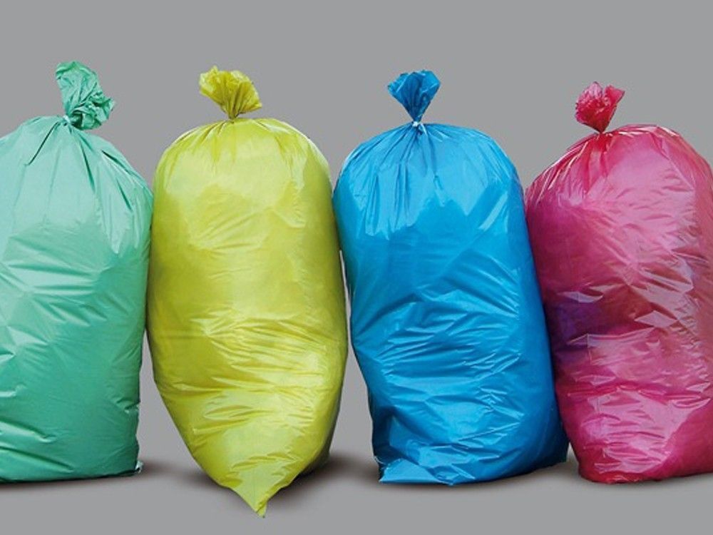 Vier farbige Plastiktüten sind aufgereiht, jede in einer anderen Farbe: grün, gelb, blau und pink. Sie sind leer und haben Griffe oben.