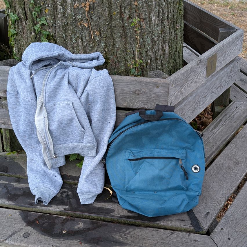 Eine graue Kapuzenjacke und ein blauer Rucksack liegen auf einer Holzbank in der Nähe eines Baumstamms.