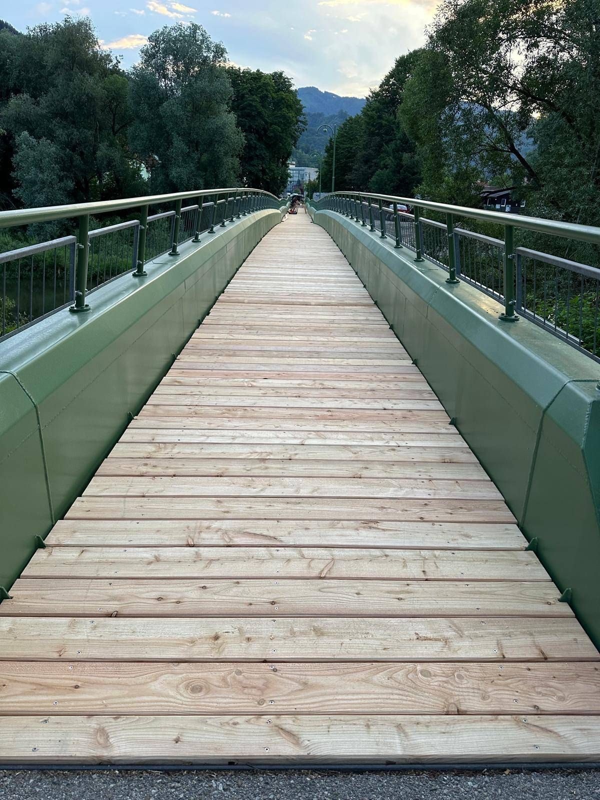 Eine hölzerne Brücke mit Metallgeländern, die sich über eine Wasserfläche erstreckt, umgeben von üppigem Grün und Gebäuden in der Ferne.