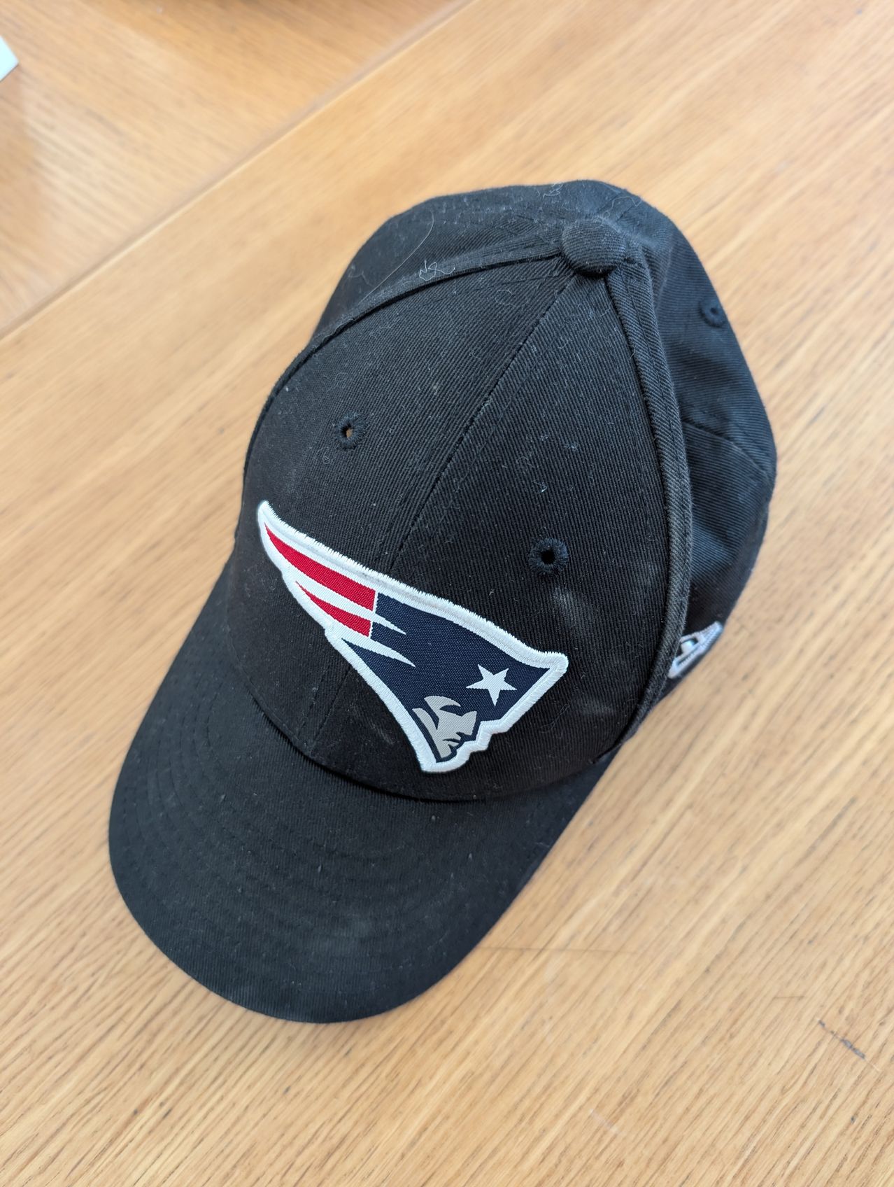Eine schwarze New England Patriots Baseball-Kappe mit einem weißen Logo auf der Vorderseite liegt auf einer Holzoberfläche.