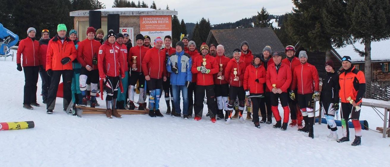 Eine Gruppe von Menschen in roter Winterkleidung steht in einem verschneiten Bereich und posiert für ein Foto. Sie halten Trophäen in den Händen und einige tragen Mützen. Hinter ihnen befindet sich ein Gebäude mit einem Schild, das Fensterhaus Johann Reith besagt.