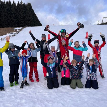 Eine Gruppe von Kindern und Erwachsenen posiert für ein Foto auf einem verschneiten Hang, trägt Winterkleidung und hält Medaillen. Einige stehen, andere knien.