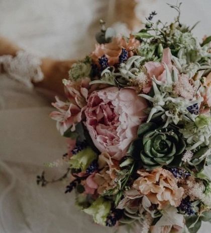 Eine Braut hält einen Blumenstrauß mit einer Mischung aus rosa, pfirsichfarbenen und grünen Blumen und Sukkulenten.