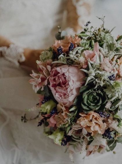 Eine Braut hält einen Blumenstrauß mit einer Mischung aus rosa, pfirsichfarbenen und grünen Blumen und Sukkulenten.
